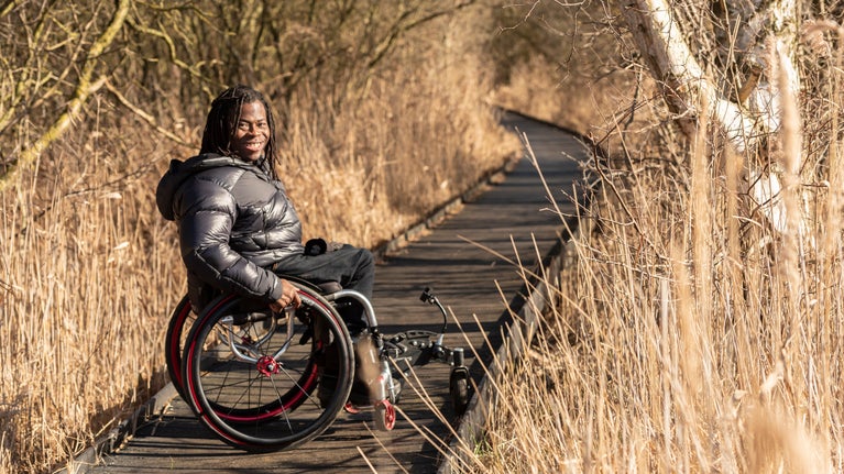 Ade Adepitan sat in his wheelchair, smiling, on the boardwalk at Wicken Fen